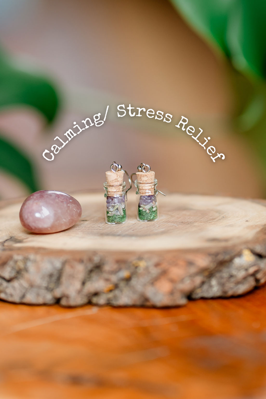 A pair of spell jar earrings with herbs and crystals inside, displayed on a wooden platform with the words 'Calming/ Stress Relief' above them.