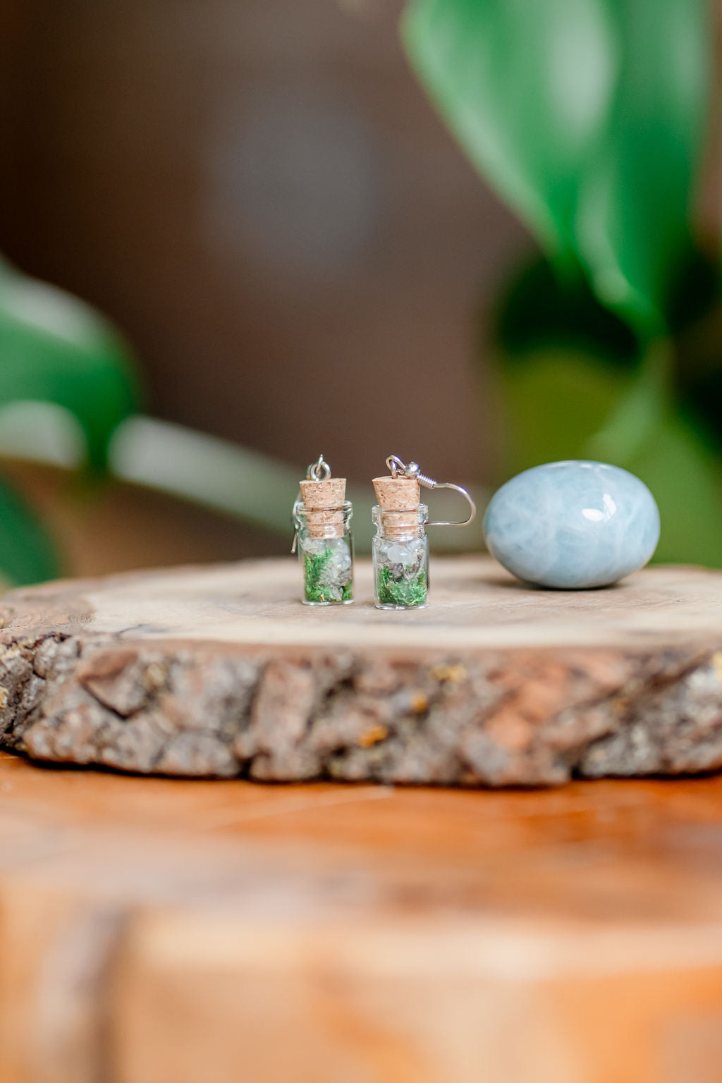 A pair of spell jar earrings made with herbs and crystals displayed on a wooden platform with a blurred green plant in the background.