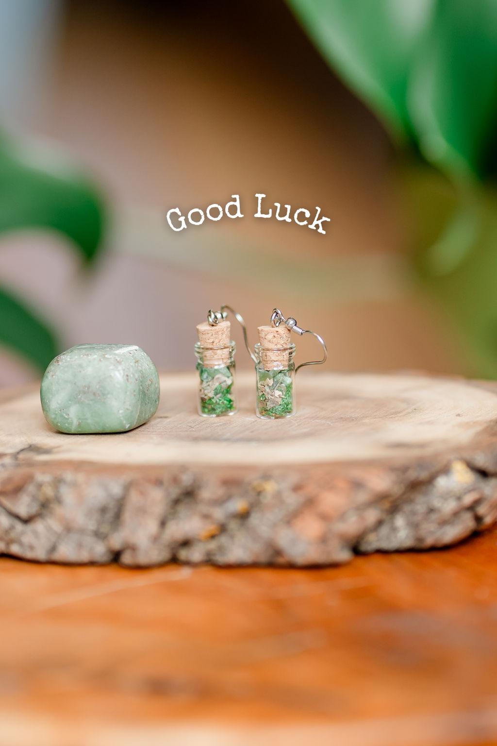 A pair of spell jar earrings with herbs and crystals displayed on a wooden platform with the text 'Good Luck' above them.