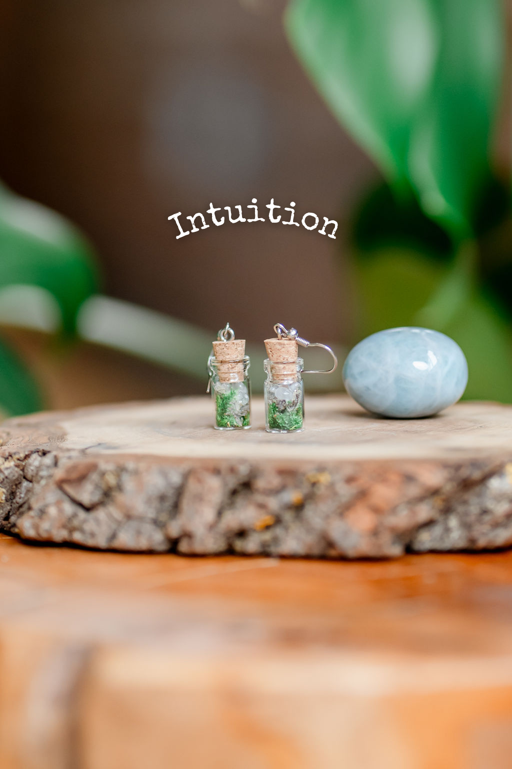 A pair of earrings designed as spell jars with herbs and crystals inside, placed on a wooden surface, with the word 'Intuition' above them and a blue crystal to the side.
