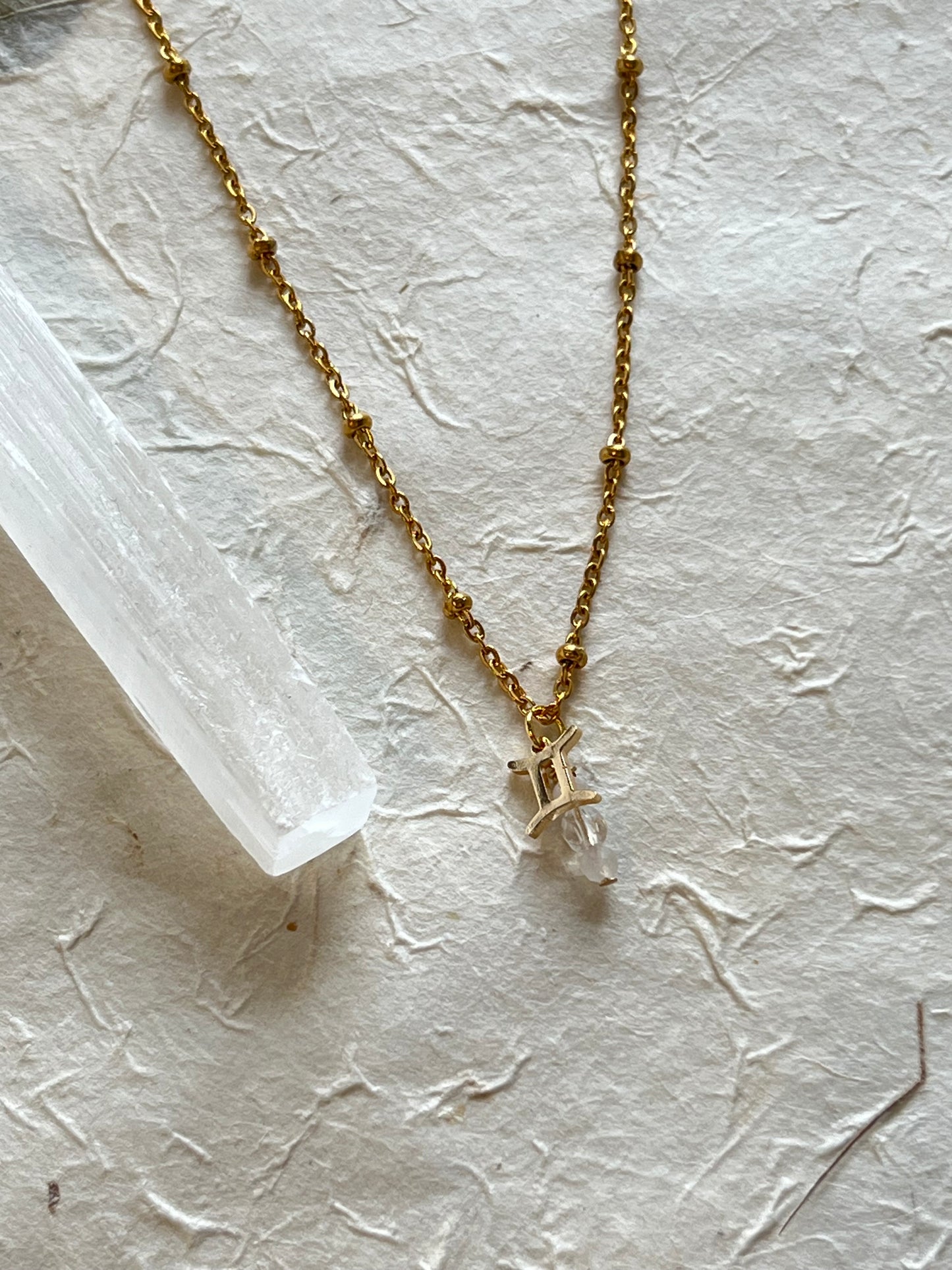 A gold necklace with a zodiac sign charm and white moonstone and clear quartz pendants, placed on a textured white surface.
