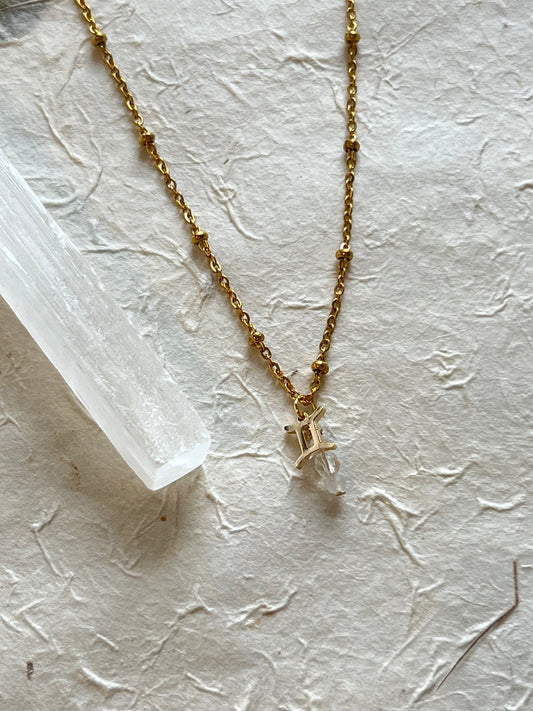 A gold necklace with a zodiac sign charm and white moonstone and clear quartz pendants, placed on a textured white surface.
